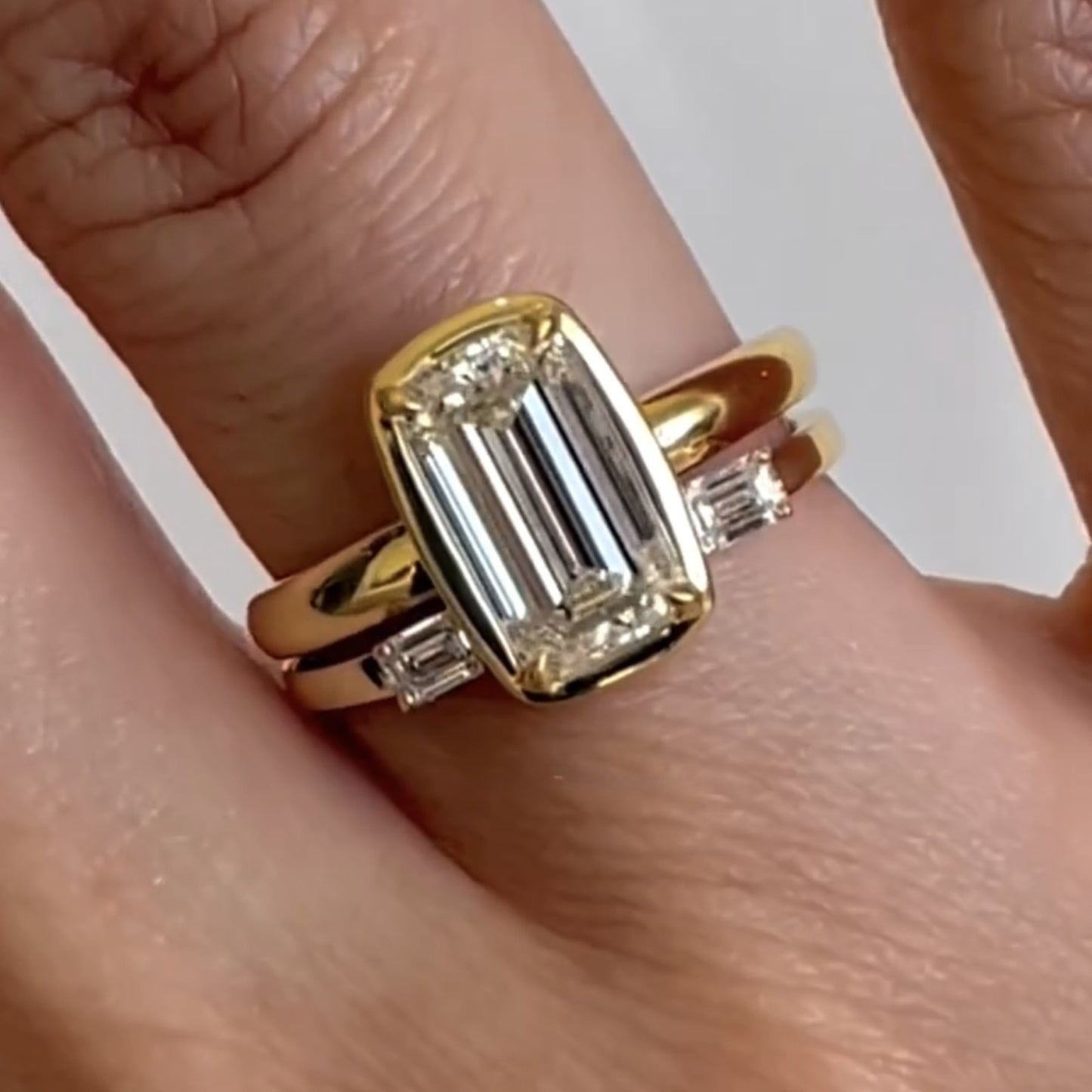 gold ring with emerald-cut diamond on a finger against a neutral background