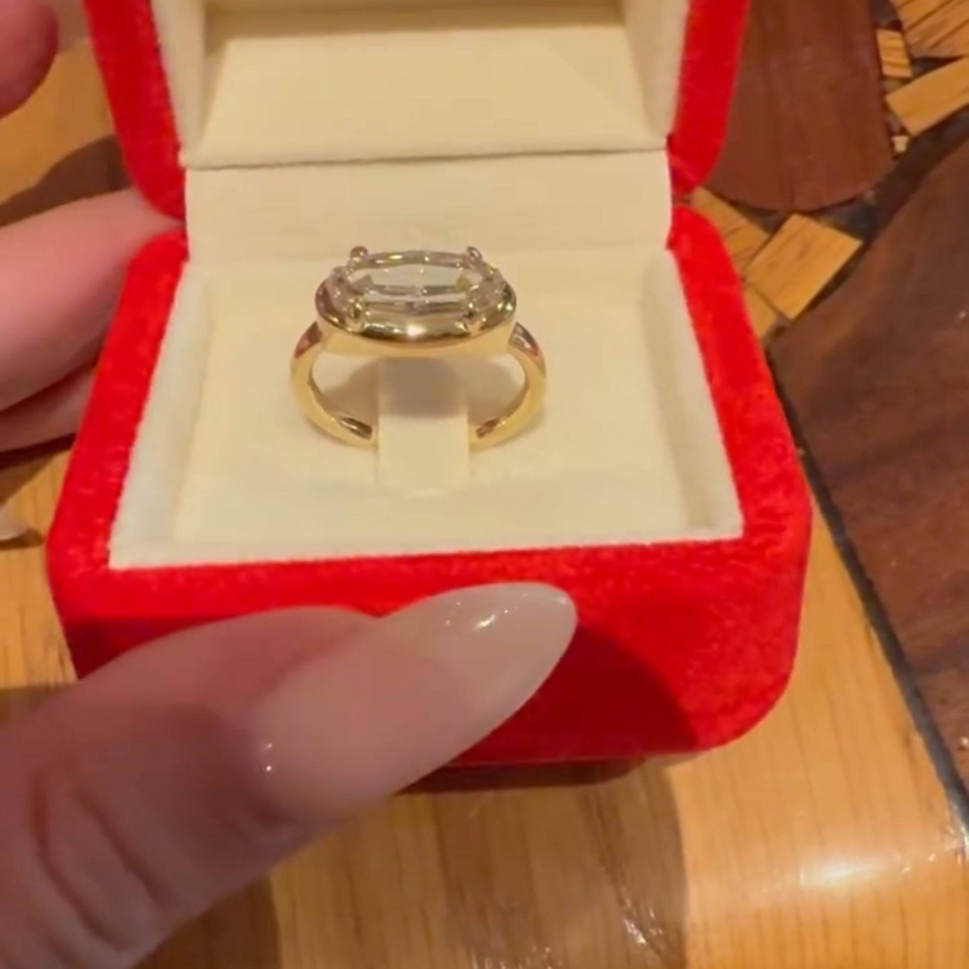 Gold ring with a large diamond in a red box held by a hand.
